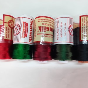 Puede incluir: Cinco carretes de hilo para bordar en rojo, verde, granate, verde oscuro y negro. Los carretes están etiquetados con "Sunsilk", "Unsilk" y "Komal Textiles".