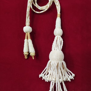 Può includere: Una collana di perle bianche con un cordoncino dorato. Il fiocco è composto da più strati di perle e ha una perla rotonda in cima.