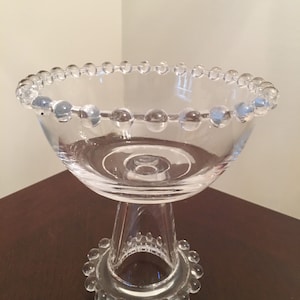 Puede incluir: Un cuenco de pedestal de vidrio transparente con un borde y una base con cuentas. El cuenco está sobre una superficie de madera oscura.