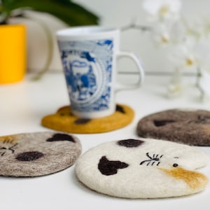 May include: Four felted wool cat-shaped coasters in brown, gray, white, and tan. The coasters are arranged on a white surface with a blue and white teacup and saucer in the background.