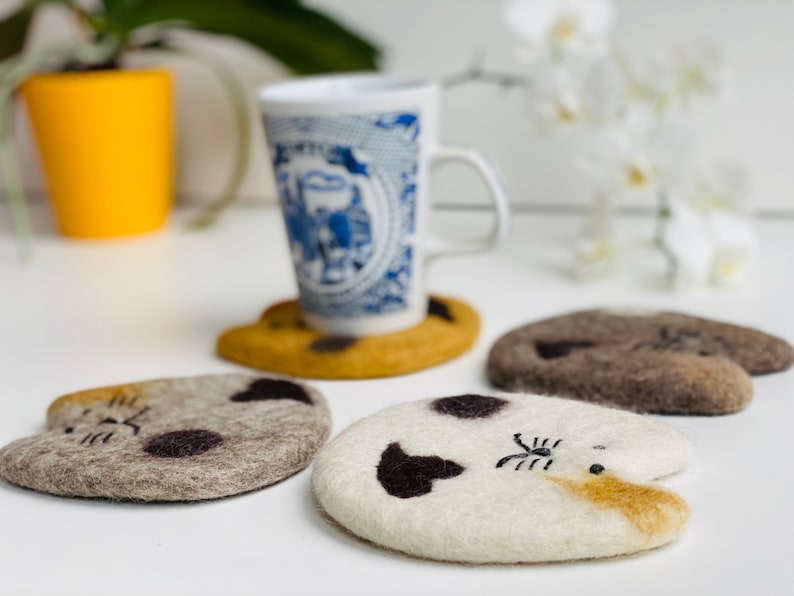 May include: Four felted wool cat-shaped coasters in brown, gray, white, and tan. The coasters are arranged on a white surface with a blue and white teacup and saucer in the background.