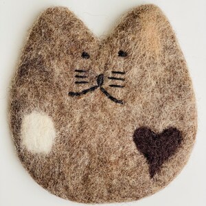 May include: A brown felt cat with a white spot and a brown heart. The cat has black embroidered eyes and whiskers.