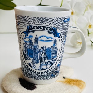 May include: White ceramic mug with a blue and white design featuring the text "BOSTON MASSACHUSETTS" and a depiction of the Boston skyline. The mug is sitting on a white and brown felted coaster.