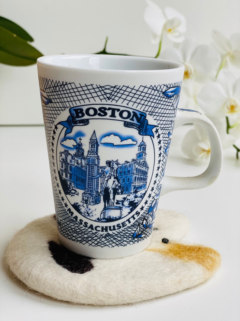 May include: White ceramic mug with a blue and white design featuring the text "BOSTON MASSACHUSETTS" and a depiction of the Boston skyline. The mug is sitting on a white and brown felted coaster.