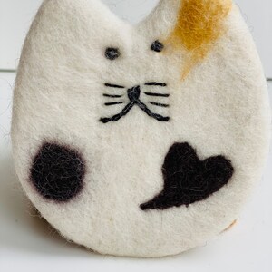 May include: White felt cat with black embroidered eyes, whiskers, and a heart shape. The cat has a black spot on one side and a tan spot on the other.