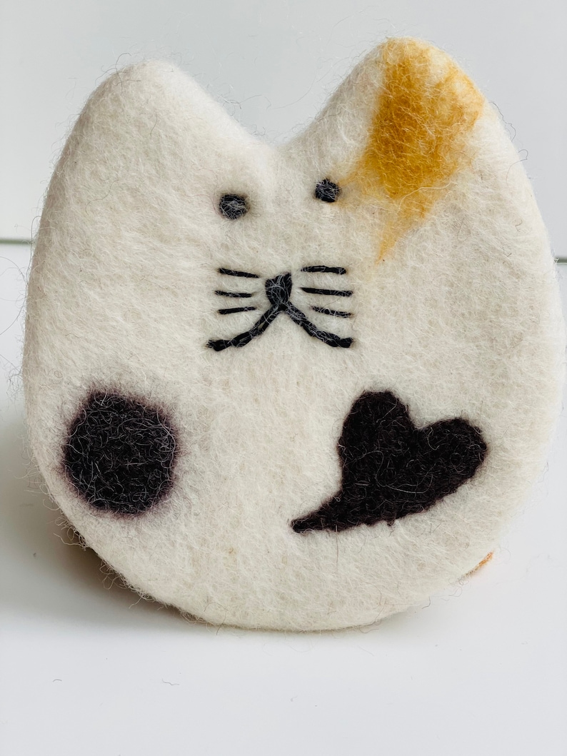 May include: White felt cat with black embroidered eyes, whiskers, and a heart shape. The cat has a black spot on one side and a tan spot on the other.