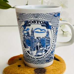May include: White ceramic mug with a blue and white illustration of the Boston skyline. The mug says "BOSTON MASSACHUSETTS" and has a black and white grid pattern around the image. The mug is sitting on a brown felt coaster.