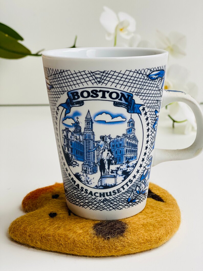 May include: White ceramic mug with a blue and white illustration of the Boston skyline. The mug says "BOSTON MASSACHUSETTS" and has a black and white grid pattern around the image. The mug is sitting on a brown felt coaster.