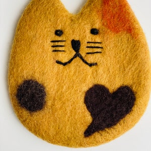 May include: A brown felt cat with black eyes, nose, whiskers, and paw prints. The cat has a black felt heart on its chest and a small orange patch on its back.