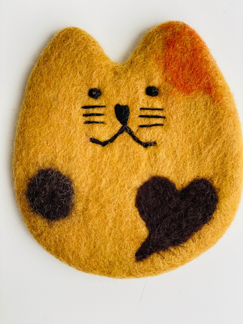 May include: A brown felt cat with black eyes, nose, whiskers, and paw prints. The cat has a black felt heart on its chest and a small orange patch on its back.