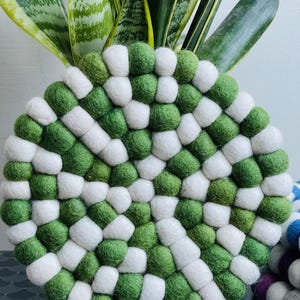 May include: A round trivet with a checkerboard design made from green and white felt balls. This handmade trivet is designed to protect surfaces from heat. The trivet is approximately 20 cm in diameter, suitable for hot pots and pans.