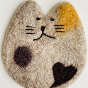 May include: A felted cat-shaped coaster with a brown, black, and yellow design. The coaster has a black embroidered face with whiskers and a heart-shaped black spot.