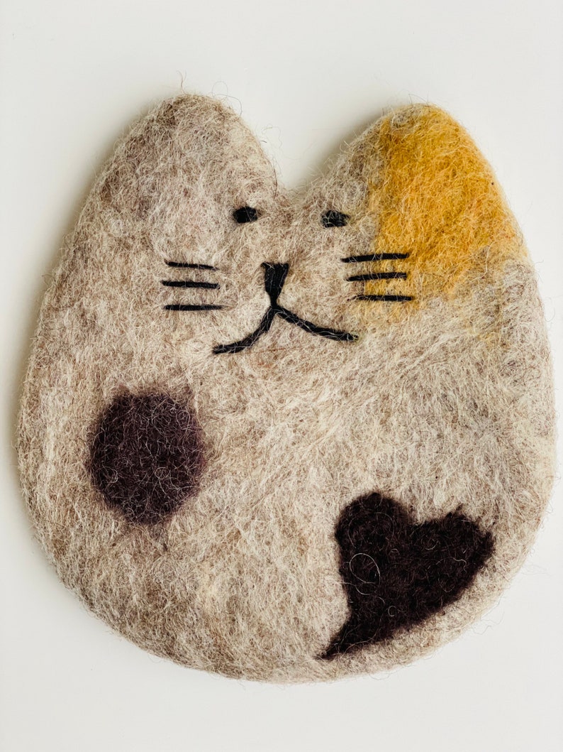 May include: A felted cat-shaped coaster with a brown, black, and yellow design. The coaster has a black embroidered face with whiskers and a heart-shaped black spot.