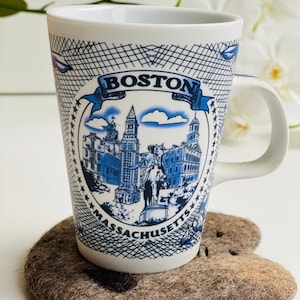 May include: White ceramic mug with a blue and white design featuring the Boston skyline and the text "BOSTON MASSACHUSETTS". The mug is sitting on a brown felt coaster.