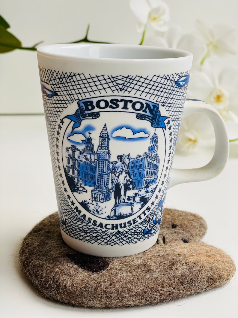 May include: White ceramic mug with a blue and white design featuring the Boston skyline and the text "BOSTON MASSACHUSETTS". The mug is sitting on a brown felt coaster.