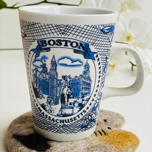 May include: A white ceramic mug with a blue and white design featuring the Boston skyline and the text "BOSTON MASSACHUSETTS". The mug is sitting on a gray felt coaster with a brown cat design.