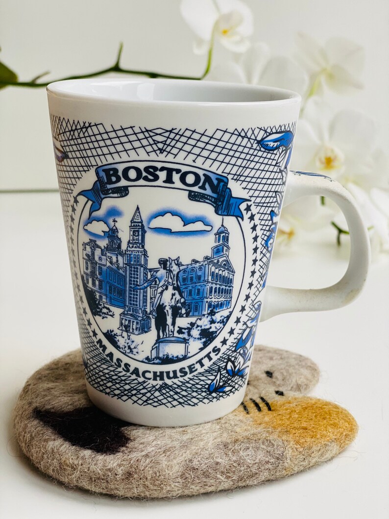 May include: A white ceramic mug with a blue and white design featuring the Boston skyline and the text "BOSTON MASSACHUSETTS". The mug is sitting on a gray felt coaster with a brown cat design.