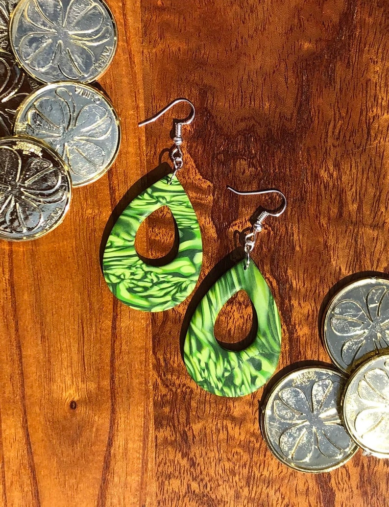 May include: A pair of green and white dangle earrings with a teardrop shape and a hole in the centre. The earrings are made of polymer clay and have a marbled effect.