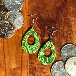 May include: A pair of green and black dangle earrings with a teardrop shape and a cutout in the centre. The earrings are on a wooden surface with gold coins.