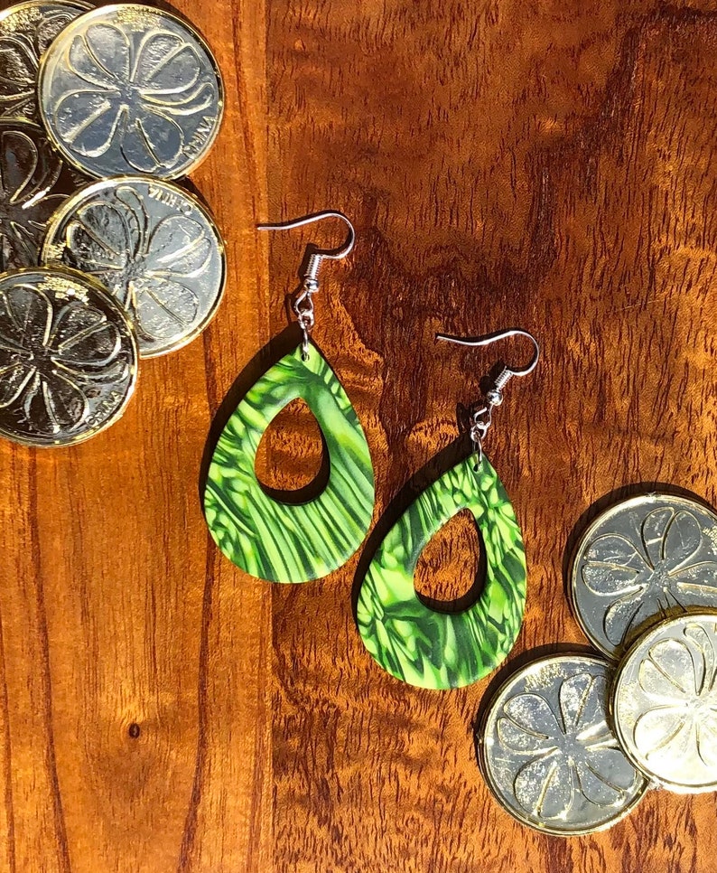 May include: A pair of green and black dangle earrings with a teardrop shape and a cutout in the centre. The earrings are on a wooden surface with gold coins.