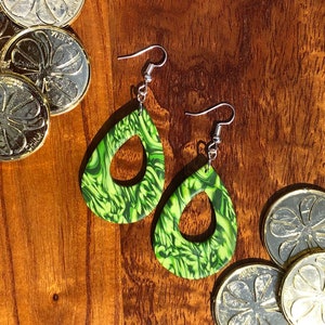 May include: A pair of green and black marbled teardrop-shaped earrings with silver hooks. The earrings have a swirl pattern and a hole in the centre.