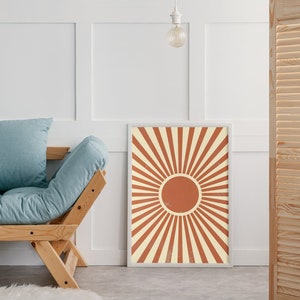 Op de afbeelding: Een ingelijste print van een gestileerde zon met oranje en witte stralen. De zon is een volle oranje cirkel in het midden van de print. De print hangt aan een witte muur.