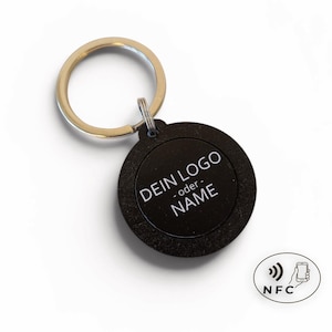 May include: Black round keychain with a gold-colored ring. The keychain has the text "DEIN LOGO - oder - NAME" in white. An NFC symbol is also present. The keychain is likely for personalization.
