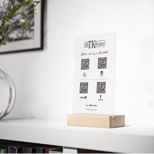 Könnte beinhalten: Ein weißes Acrylschild mit schwarzem Text und QR-Codes. Das Schild lautet "Mr. TK Booker" und "Bleib mit uns in Kontakt.". Das Schild steht auf einem Holzständer.