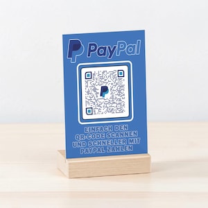 Op de afbeelding: Een blauw PayPal QR-codeschild op een houten voet. Het bord toont het PayPal-logo en een QR-code, met de tekst "Einfach den QR-Code scannen und schneller mit PayPal zahlen."
