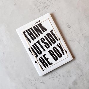 Könnte beinhalten: Ein rechteckiges weißes Keramiktablett mit einem erhabenen weißen Rand und fettgedrucktem schwarzem Text, der THINK OUTSIDE THE BOX lautet. Das Design ist auf der flachen Oberfläche zentriert und verleiht diesem Dekorationsgegenstand ein klares Aussehen.
