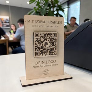 Könnte beinhalten: Ein hölzerner Tischaufsteller mit einem QR-Code für PayPal-Zahlungen. Der Text auf dem Aufsteller lautet "MIT PAYPAL BEZAHLEN" und "DEIN LOGO". Der Aufsteller ist hellbraun und hat eine rechteckige Form.