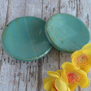May include: Two teal ceramic dishes with a gold glaze, sitting on a white wooden surface, with three yellow flowers with red centres.