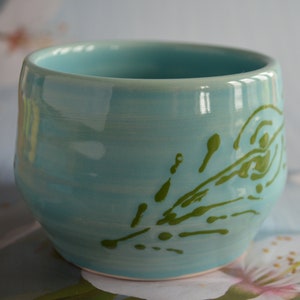 May include: A light blue ceramic mug with a green abstract design. The mug is sitting on a white surface with a pink flower.