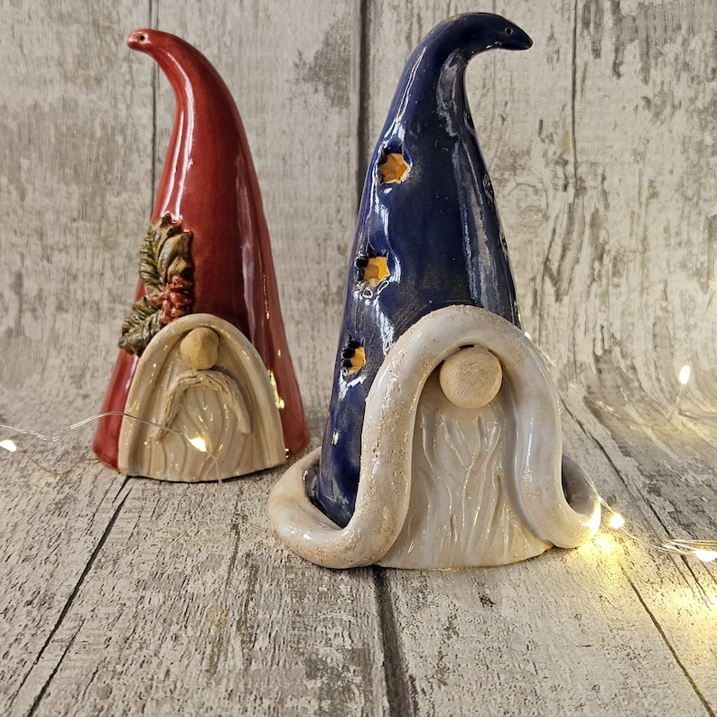 Pu&ograve; includere: Due statuette di gnomi in ceramica. Una &egrave; rossa con un motivo decorativo di foglie e bacche, l'altra &egrave; blu scuro con ritagli a forma di stella. Entrambi hanno barbe bianche e cappelli con punte curve. Gli gnomi sono su una superficie di legno.
