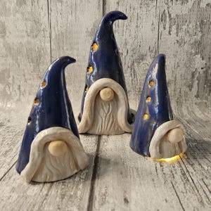 Pu&ograve; includere: Tre statuette di gnomi in ceramica con cappelli a punta blu scuro e barbe color crema. I cappelli hanno piccoli ritagli rotondi che brillano di una luce calda. Gli gnomi sono disposti su una superficie di legno grigio invecchiato.
