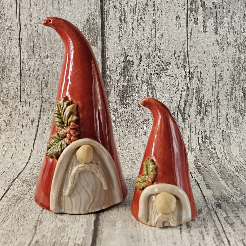Pu&ograve; includere: Due statuette di gnomi in ceramica. Gli gnomi hanno alti cappelli a punta rossi e barbe bianche. Uno gnomo &egrave; pi&ugrave; grande dell'altro. I cappelli hanno una finitura lucida e sono decorati con foglie e bacche verdi e marroni.
