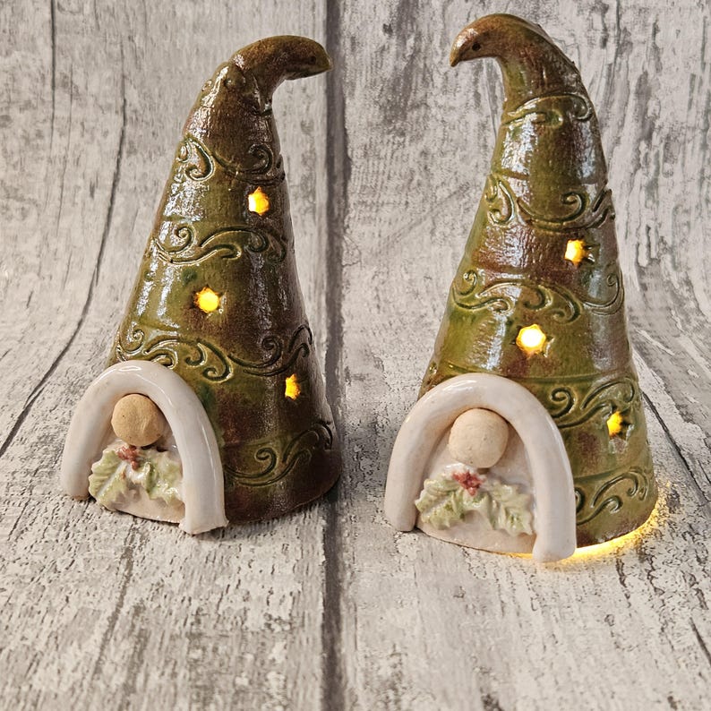 Pu&ograve; includere: Due statuette di gnomi in ceramica con cappelli conici smaltati verdi e marroni. I cappelli hanno volute decorative e ritagli a forma di stella che si illuminano. Ogni gnomo ha una base bianca ad arco con una piccola figura e foglie di agrifoglio.