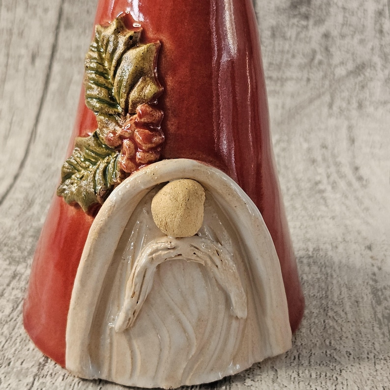 Pu&ograve; includere: Statuina di gnomo in ceramica rossa con barba bianca e testa beige. Lo gnomo ha un motivo decorativo di foglie e pigne sul lato. La statuina &egrave; su una superficie di legno.
