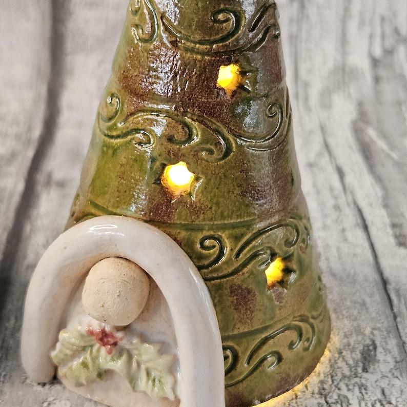 Pu&ograve; includere: Un portacandele a forma di albero di Natale in ceramica con una glassa verde e marrone. L'albero ha ritagli a forma di stella che emettono un bagliore caldo. Un arco bianco incornicia una piccola figura con agrifoglio.