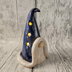 Pu&ograve; includere: Statuetta di mago in ceramica con un cappello blu scuro con tre ritagli a forma di stella che si illuminano di giallo. La statuetta ha una barba bianca e un bordo bianco attorno alla base del cappello. Sfondo in legno grigio chiaro.
