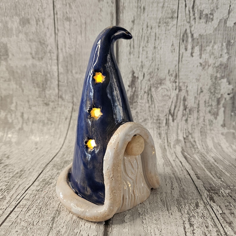 Pu&ograve; includere: Statuetta di mago in ceramica con un cappello blu scuro con tre ritagli a forma di stella che si illuminano di giallo. La statuetta ha una barba bianca e un bordo bianco attorno alla base del cappello. Sfondo in legno grigio chiaro.