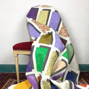 May include: A colorful patchwork quilt with a geometric design featuring squares and rectangles in shades of purple, green, yellow, orange, blue, and brown. The quilt is draped over a chair with a red velvet seat.