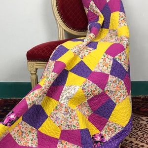 K&ouml;nnte beinhalten: Eine gelbe, violette und rosafarbene Patchwork-Decke mit Blumenmuster liegt &uuml;ber einem roten Samtstuhl. Die Decke hat einen gelben Rand.