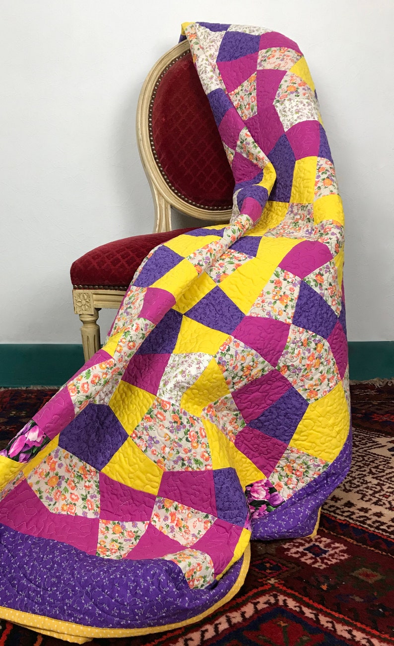 K&ouml;nnte beinhalten: Eine gelbe, violette und rosafarbene Patchwork-Decke mit Blumenmuster liegt &uuml;ber einem roten Samtstuhl. Die Decke hat einen gelben Rand.