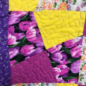 K&ouml;nnte beinhalten: Ein Patchwork-Deckenst&uuml;ck mit verschiedenen Blumenmustern in Violett-, Rosa-, Gelb- und Wei&szlig;t&ouml;nen. Die Decke besteht aus verschiedenen Stoffen und ist mit einem wellenf&ouml;rmigen Stich gesteppt.