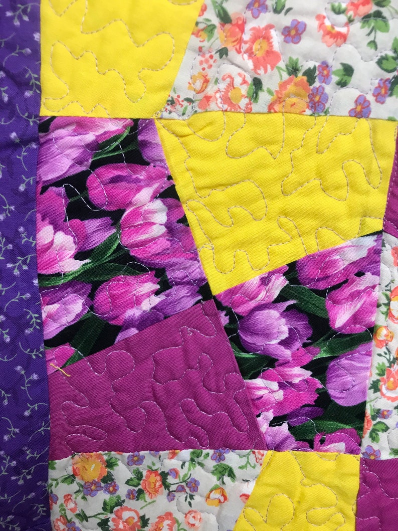 K&ouml;nnte beinhalten: Ein Patchwork-Deckenst&uuml;ck mit verschiedenen Blumenmustern in Violett-, Rosa-, Gelb- und Wei&szlig;t&ouml;nen. Die Decke besteht aus verschiedenen Stoffen und ist mit einem wellenf&ouml;rmigen Stich gesteppt.