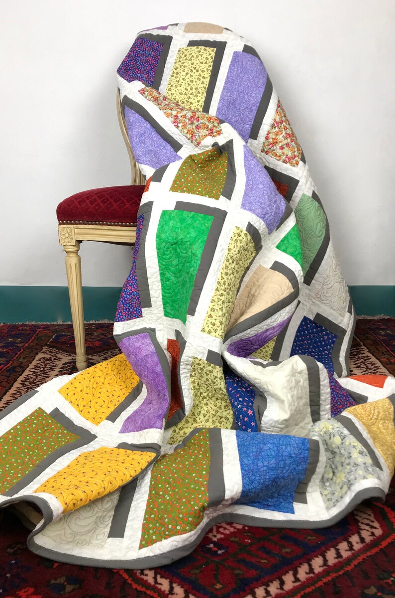 May include: A colorful patchwork quilt with a geometric design featuring squares and rectangles in shades of purple, green, yellow, orange, blue, and brown. The quilt is draped over a chair with a red velvet seat.