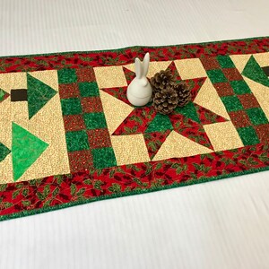 Pode incluir: Um corredor de mesa acolchoado com tema natalino. Apresenta um esquema de cores vermelho, verde e creme com desenhos de estrelas, árvores e folhas de azevinho. O corredor de mesa mede aproximadamente 137 cm de comprimento.