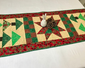 May include: A quilted table runner with a Christmas theme. It features a red, green and cream colour scheme with a star, trees and holly leaf designs. The runner is approximately 54 inches long.