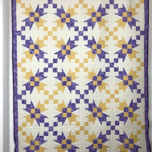 Peut inclure: Une grande courtepointe avec un motif d'étoiles répétitif dans des tons dorés et violets sur un fond crème. La courtepointe a une bordure violette et est suspendue à une tringle en métal. Le motif en étoile est composé de carrés et de triangles.